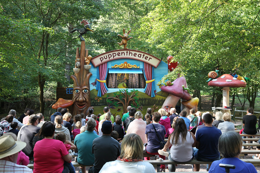 Erwachsene und Kinder blicken auf dei Bühne eines Puppentheaters umgeben von Wald.